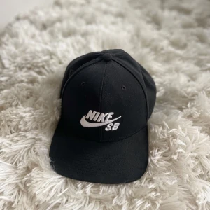 Svart keps från Nike SB - Snygg svart keps från Nike SB med broderad logga framtill. Kepsen har en klassisk design med justerbar rem baktill för perfekt passform. Liten flaw i loggan då en tråd är lös, går säkert att ta bort. 