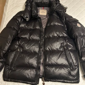 Svart dunjacka från Moncler - Säljer en snygg svart dunjacka från Moncler med dragkedja och hög krage. Jackan har en quiltad design och två praktiska fickor med dragkedjor. Perfekt för kalla dagar när du vill hålla stilen. Passar storlek L, Är 180 och väger 75kg passade perfekt. Den är i perfekt skick har bara haft den i nån månad 