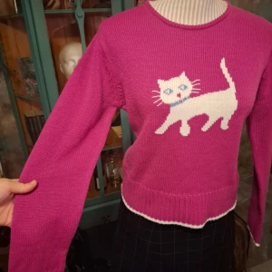 Rosa stickad tröja med kattmotiv från Select - Charmig rosa stickad tröja från Select med ett sött vitt kattmotiv på framsidan. Tröjan har långa ärmar och en rund halsringning. Perfekt för att ge en lekfull touch till din outfit!