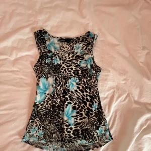 Mönstrad ärmlös topp från KappAhl - Snygg ärmlös topp från KappAhl med ett djärvt leopardmönster i svart och vitt, kombinerat med stora blå blommor. Toppen har dekorativa spännen på axlarna som ger en extra touch. Perfekt för att liva upp garderoben! Passar allt från s till l