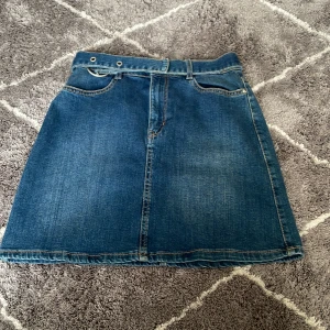 Jeanskjol från Lindex - Snygg jeanskjol från Lindex i klassisk blå denim. Kjolen har en kort längd med två fickor bak och en justerbar rem med metallringar i midjan. Perfekt för en avslappnad stil.