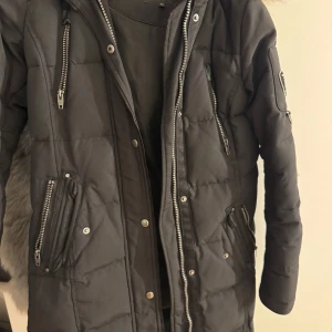 Svart dunjacka med pälsdetaljer från Rockandblue - Snygg svart dunjacka från Rockandblue med pälsdetaljer på huvan. Jackan har flera dragkedjor och en cool patch på ärmen. Perfekt för kalla dagar med sin quiltade design och långa ärmar.