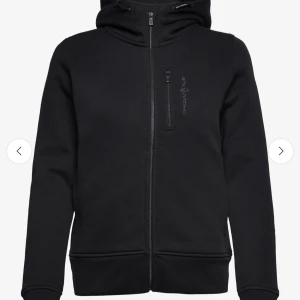 Svart hoodie från Sail Racing - Snygg svart hoodie från Sail Racing med dragkedja framtill och en praktisk bröstficka med dragkedja. Perfekt för kyliga dagar med sin bekväma passform och huva.