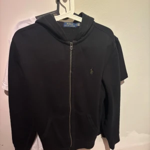 Svart hoodie från Polo Ralph Lauren - Säljer en svart hoodie från Polo Ralph Lauren med dragkedja och huva. Den har långa ärmar och en liten logga på bröstet. Perfekt för en avslappnad stil.