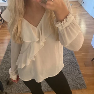 Vit blus med volangdetaljer från Zara - Säljer en elegant vit blus med v-ringning och volangdetaljer framtill. Perfekt för både vardag och speciella tillfällen. Har aldrig använt den så den är som ny