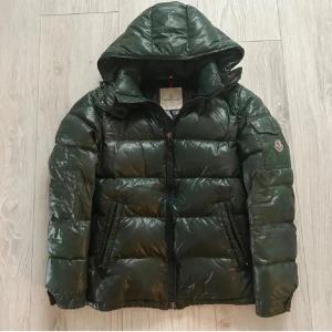 Grön dunjacka från Moncler - Säljer en snygg grön dunjacka från Moncler med huva och dragkedja. Jackan har en glansig finish och är perfekt för kyligare väder. Den har långa ärmar och två fickor framtill. Litet hål på armen som är igensydd men det är inget som syns! Jackan har även lite regnskada! Tyvärr har själva saken som man drar upp dragkedjan med åkt av men det går fortfarande att stänga och öppna och är lätt att köpa en ny! 