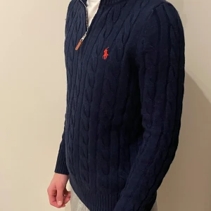 Ralph Lauren stickad halfzip  - Säljer en stilren mörkblå stickad tröja från Ralph Lauren med dragkedja och det klassiska röda logotypbroderiet på bröstet. Tröjan har en kabelstickad design och långa ärmar, perfekt för kyligare dagar. Passar både till vardags och mer uppklädda tillfällen. Nyskick, använd 1 gång❗️