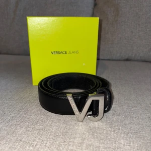 Svart bälte från Versace Jeans - Snyggt svart bälte från Versace Jeans med en stilren silverfärgad spänne i form av bokstäverna VJ. Tillverkat i skinn och kommer i originalförpackning. Perfekt för att ge en lyxig touch till din outfit.                                                           Storlek 100/115.                                                                  Nypris (850kr)