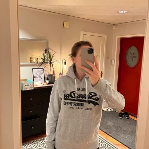 Grå hoodie från Superdry - Säljer en grå hoodie från Superdry med svart tryck på framsidan. 