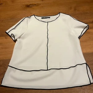 Vit blus från Zara Basic Collection - Säljer en stilren vit blus från Zara Basic Collection med svarta kantdetaljer. Blusen har korta ärmar och en rund halsringning. Perfekt för en minimalistisk look.