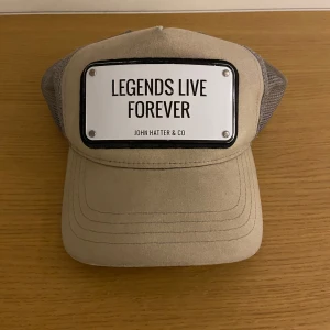 Beige keps från John Hatter & Co - Snygg beige keps med texten 'Legends Live Forever' på en vit platta framtill. Kepsen är ändå bra skick och sitter bra på huvudet, de ändå tråkiga är att plåten släppt i ena kanten (se sista bild). Men vill man ha en snygg o bekväm keps för pengarna så är de bara att höra av sig!