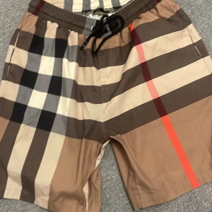 Rutig badshorts från Burberry - Snygga badshorts från Burberry med klassiskt rutigt mönster i beige, svart och röd. De har en elastisk midja med snörning för perfekt passform. Perfekta för sommarens stranddagar!