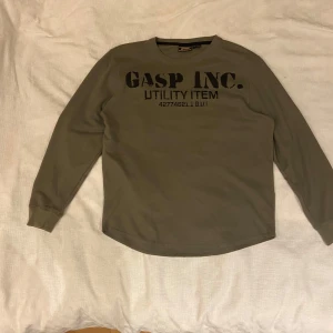 Grå långärmad tröja från GASP - Säljer en grå långärmad tröja från GASP med texten 'GASP INC. UTILITY ITEM' tryckt på framsidan. Tröjan har en ribbad struktur och rund halsringning. Perfekt för gymmet.