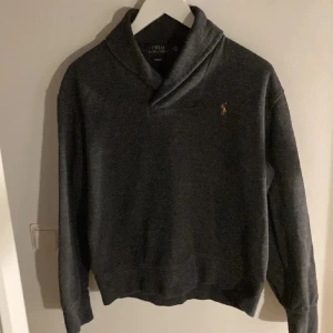  Ralph Lauren Tröja  - Säljer en stilren grå tröja från Ralph Lauren med en liten broderad logga på bröstet. Tröjan har långa ärmar och en unik krage som ger en elegant touch. Perfekt för kyligare dagar.