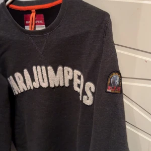 Mörkgrå sweatshirt från Parajumpers - Säljer en mörkgrå sweatshirt från Parajumpers med stort vitt broderat märke på bröstet och en patch på ärmen. Tröjan har långa ärmar och en rund halsringning. Perfekt för en avslappnad stil.