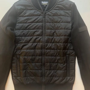 Barbour Hybrid Zip Up - Svart quiltad jacka från Barbour med dragkedja framtill. Jackan har långa ärmar och en ribbad krage. 