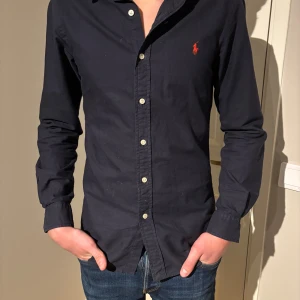 Svart långärmad skjorta från Ralph Lauren - Snygg svart skjorta från Ralph Lauren med klassisk krage och knappar framtill. Den har en broderad röd logga på bröstet och långa ärmar. Perfekt för en stilren look. Slim fit. 