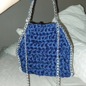 Blå virkad väska med kedjehandtag - Snygg blå virkad väska med silverfärgade kedjehandtag. Perfekt för att ge din outfit en unik touch. Väskan har en mjuk och texturerad yta som ger en cool look.