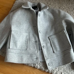 Grå kappa från Uniqlo - Snygg grå kappa från Lager 157 med en stilren design. Jackan har stora fickor framtill och knäpps med knappar. Perfekt för kyligare dagar med sin klassiska look.