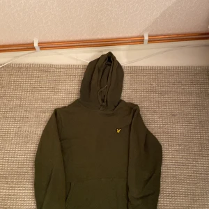 Olivgrön hoodie från Lyle & Scott - Säljer en olivgrön hoodie från Lyle & Scott med en liten gul logga på bröstet. Tröjan har en klassisk design med huva och långa ärmar. Perfekt för en avslappnad stil.