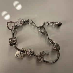 Silverfärgat armband med hjärtberlocker - Charmigt silverfärgat armband med flera hjärtformade berlocker. Berlockerna är dekorerade med glittriga stenar och rosa detaljer. Armbandet har en justerbar kedja med ett litet hjärta i slutet. Det är jätte lite slitet men inget man ser.