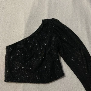 Svart paljettprydd oneshoulder topp från Lycilia - Snygg svart oneshoulder topp från Lycilia, täckt med glittrande paljetter. Perfekt för en kväll ute eller en fest. Toppen har en lång ärm med puff och en elegant passform som framhäver axeln.