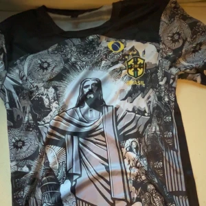 Fotbollströja med Brasilien-tema - Unik fotbollströja med ett detaljerat tryck av Kristusstatyn och CBF-logga. Tröjan har korta ärmar och en mix av svart, grå och vita nyanser. Perfekt för den som vill sticka ut på läktaren eller planen.