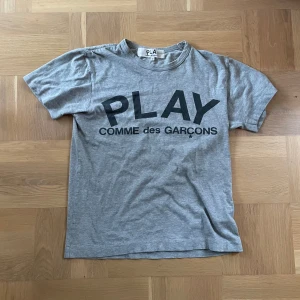 Grå t-shirt från Comme des Garçons - Säljer en grå t-shirt från Comme des Garçons med trycket 'PLAY' på framsidan. T-shirten är kortärmad och har en klassisk rund halsringning. Perfekt för en avslappnad stil.