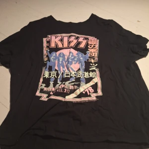 Svart KISS t-shirt med japanskt tryck - Cool svart t-shirt med KISS-tryck och japansk text. Perfekt för fans av bandet! T-shirten har korta ärmar och är gjord i bomull för en bekväm passform.