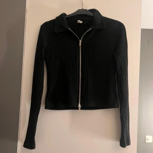 Svart ribbad tröja med dragkedja - Snygg svart ribbad tröja med dragkedja framtill och krage. Tröjan har långa ärmar och en stilren design som passar perfekt till olika tillfällen.