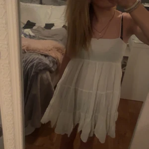 Vit sommarklänning med smala axelband - Säljer en söt vit sommarklänning med smala axelband och en luftig passform. Klänningen har en smockad överdel och faller i flera lager för en bohemisk känsla. Perfekt för varma dagar!