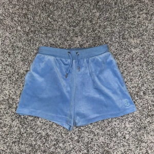 Blå velourshorts från Juicy Couture - Snygga blå velourshorts från Juicy Couture med en broderad logga på benet. De har en elastisk midja med snörning och en bakficka. Perfekta för en avslappnad stil.