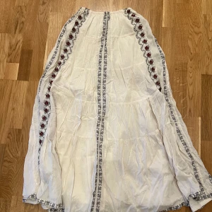 Vit klänning från Zara - Säljer en vacker vit klänning från Zara med broderade detaljer i rött och svart längs sidorna. Klänningen har en luftig passform och är perfekt för varma dagar. Den är tillverkad i ett lätt och bekvämt material. Använd 1 gång och testad