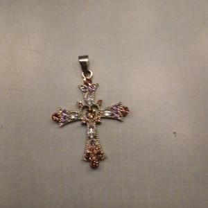 Korsformat hängsmycke med färgade stenar - Elegant korsformat hängsmycke dekorerat med färgade stenar i lila, rosa och orange nyanser. Smycket har en detaljerad design med en metallisk bas och en ögla för kedja. Perfekt för att ge en färgglad touch till din stil.