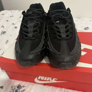 Svarta och Blåa Nike Air Max 97 - Snygga svarta Nike Air Max sneakers med blå detaljer. Skorna har en robust design med snörning och en bekväm sula. Perfekta för en sportig look.
