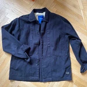 Svart jacka från Jack& Jones - Snygg svart jacka från Samsøe Samsøe med knappar framtill och två stora fickor. Jackan har en klassisk krage och är långärmad. Perfekt för en stilren look.
