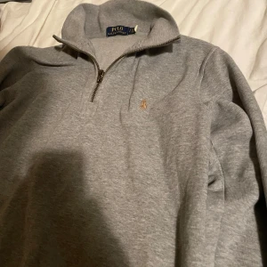 Grå tröja med dragkedja från Polo Ralph Lauren - Säljer en stilren grå tröja från Polo Ralph Lauren med halv dragkedja och broderad logga på bröstet. Perfekt för en avslappnad och snygg look. Tröjan har långa ärmar och en bekväm passform.
