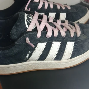 Svarta Adidas Campus sneakers - Snygga svarta Adidas Campus sneakers med vita ränder och rosa snören. Skorna har en klassisk design med Adidas-loggan på hälen och tungan. Perfekta för en avslappnad stil.