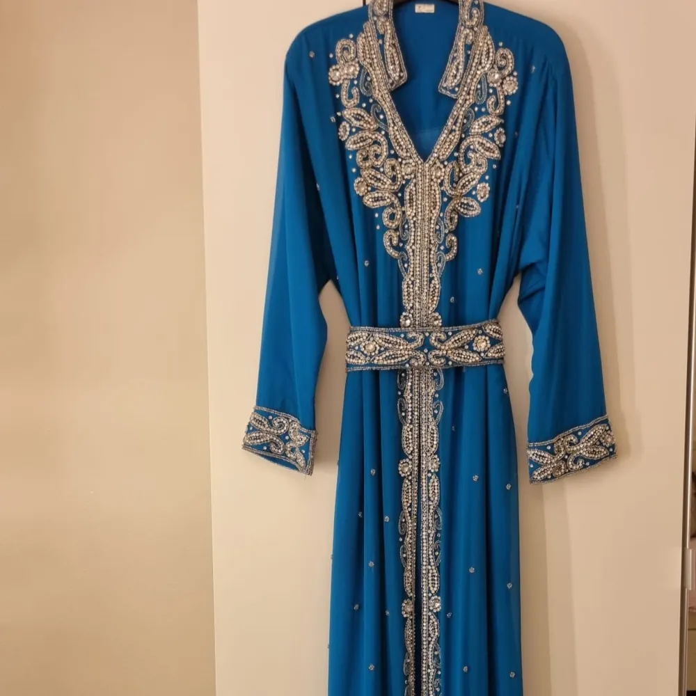 Elegant blå kaftan med detaljerade pärlbroderier längs halsringningen, ärmsluten och fållen. Klänningen har långa ärmar och ett matchande bälte i midjan för en stilfull look. Perfekt för speciella tillfällen.. Mekot.