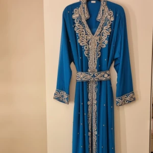 Blå kaftan med pärlor - Elegant blå kaftan med detaljerade pärlbroderier längs halsringningen, ärmsluten och fållen. Klänningen har långa ärmar och ett matchande bälte i midjan för en stilfull look. Perfekt för speciella tillfällen.