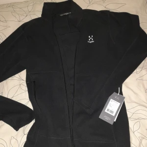 Svart fleecetröja från Haglöfs - Säljer en svart fleecetröja från Haglöfs, modell Buteo Mid Jacket. Tröjan har en dragkedja framtill och är perfekt för kyligare dagar. Perfekt för utomhusaktiviteter och vardagsbruk. Skriv vid funderingar och ge gärna BUD!