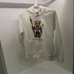 Vit hoodie från Ralph Lauren med björntryck - Säljer en vit hoodie från Ralph Lauren med ett coolt tryck av en björn i skidutrustning på framsidan. Hoodien har långa ärmar och en klassisk huva. Perfekt för en avslappnad stil med en touch av lyx.Hoodien är helt slutsåld överallt passar i storlek 14-16 år runt 160cm