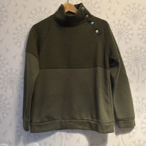 Olivgrön quiltad tröja med knappar - Snygg olivgrön tröja med quiltad design på bröstet och axlarna. Tröjan har en hög krage med dekorativa knappar på sidan. Perfekt för kyligare dagar.