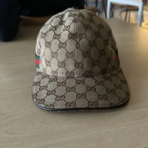 Beige keps från Gucci äkta  - Snygg beige keps från Gucci med det klassiska GG-mönstret. Kepsen har en justerbar rem baktill och detaljer i grönt och rött på sidan. Perfekt för en stilren look.