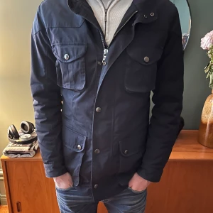 Mörkblå jacka från Hackett - Säljer en stilren mörkblå jacka från Hackett London. Jackan har flera praktiska fickor med knappar och en dragkedja framtill. Den är quiltad på insidan för extra värme och komfort. Perfekt för kyligare dagar! Ny pris 6700