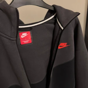 Nike tech fleece - Säljer en snygg grå och svart hoodie från Nike med röd logga på bröstet. Jackan har dragkedja och huva, perfekt för en sportig look. Den är långärmad och har praktiska fickor med dragkedjor.