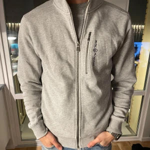 Grå hoodie från Sail Racing - Säljer en stilren grå hoodie från Sail Racing med dragkedja och en liten bröstficka. Tröjan har en bekväm passform och är perfekt för vardagsbruk. Den har en huva med logotypdetalj och långa ärmar.