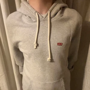 Grå hoodie från Levi's - Säljer en stilren grå hoodie från Levi's med en liten röd logga på bröstet. Tröjan har en klassisk huva med vita dragsnören och en bekväm passform. Perfekt för en avslappnad look.