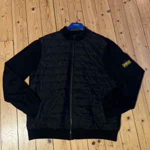 Svart jacka från Barbour International - Säljer en svart jacka från Barbour International med quiltad design och dragkedja. Jackan har långa ärmar och en ribbad krage. Perfekt för en stilren look. Storlek xl, använd mycket sparsamt. Nypris är Ca 2200 kr