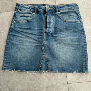 Jeanskjol från Gina Tricot - Säljer en snygg jeanskjol från Gina Tricot i klassisk blå denim. Kjolen har en knappgylf och fransig nederkant för en cool look. Två framfickor och två bakfickor. Storlek 38. 
